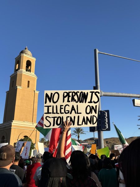 Photo courtesy of The Chaparral/Layla Freiberg. A protestor in Cathedral City holds a sign that reads "No Person is Illegal on Stolen Land." 