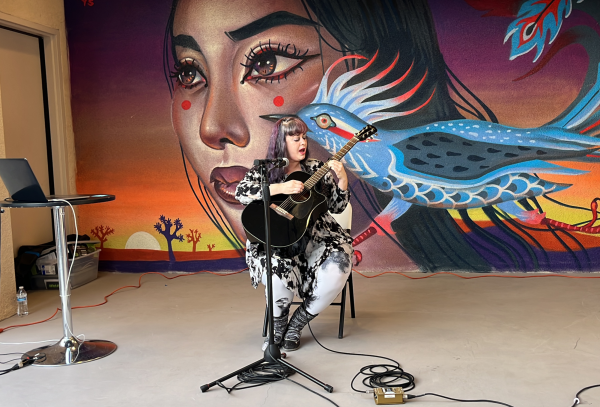 A sitting woman sings and plays acoustic guitar in front of a painted mural of a woman and a roadrunner.