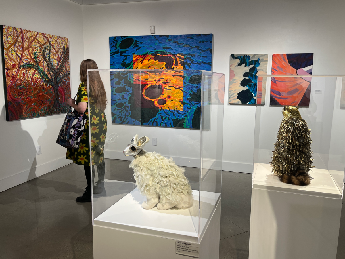 A woman stands in front of a group of large abstract paintings. In the foreground are elegant, shiny sculptures of a rabbit and a marmot.