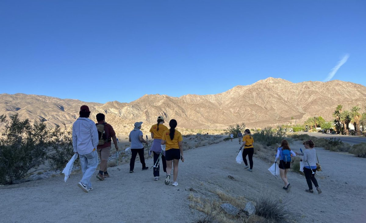 Eight peole holding trash bags and reach extenders walk into the desert. Sprawling mountains are illuminated by the morning sunlight.